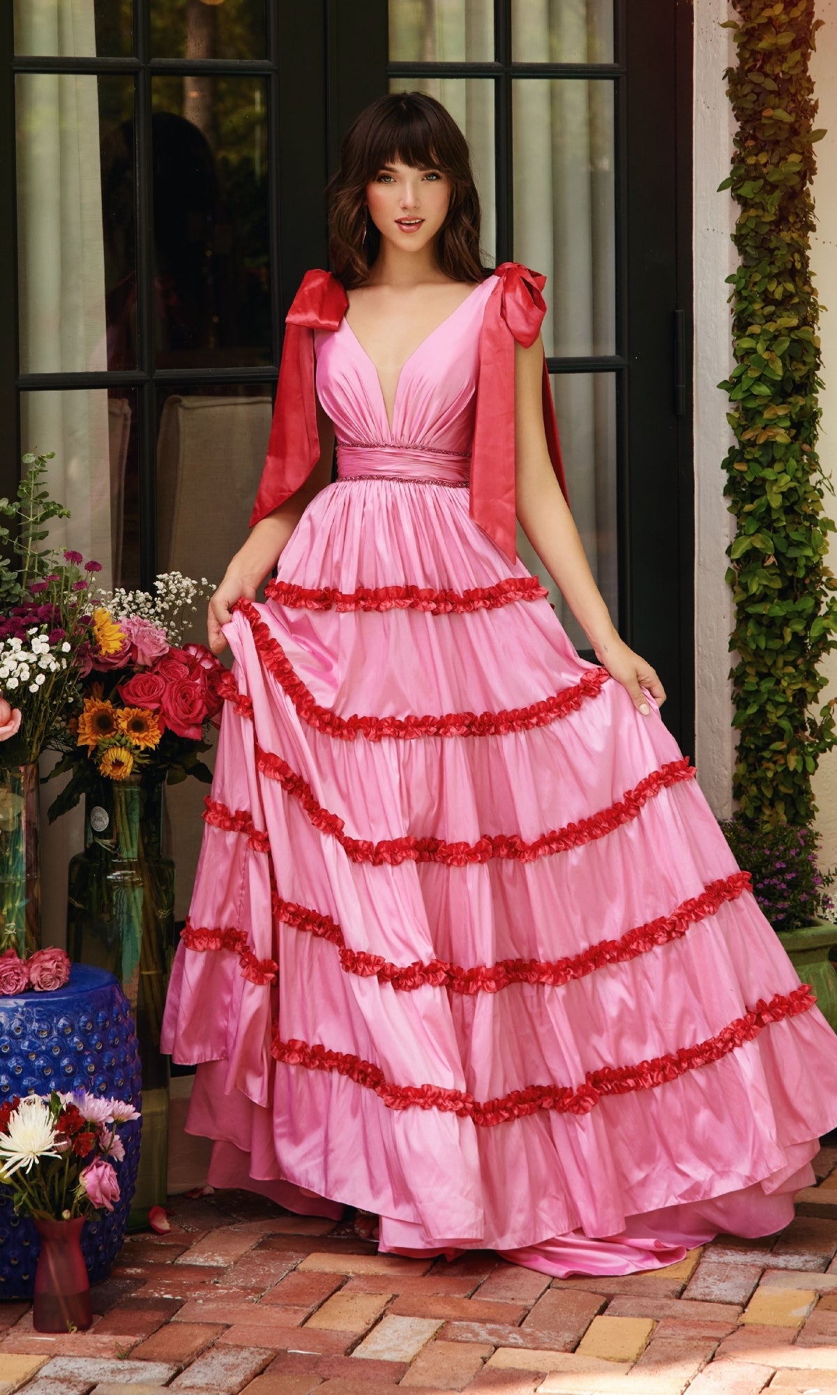 A woman stands on a brick patio in the Ava Presley 40091 Shoulder Bows A-Line Formal Dress, featuring bow tie shoulders and a v-neckline, surrounded by flower arrangements near a glass door.